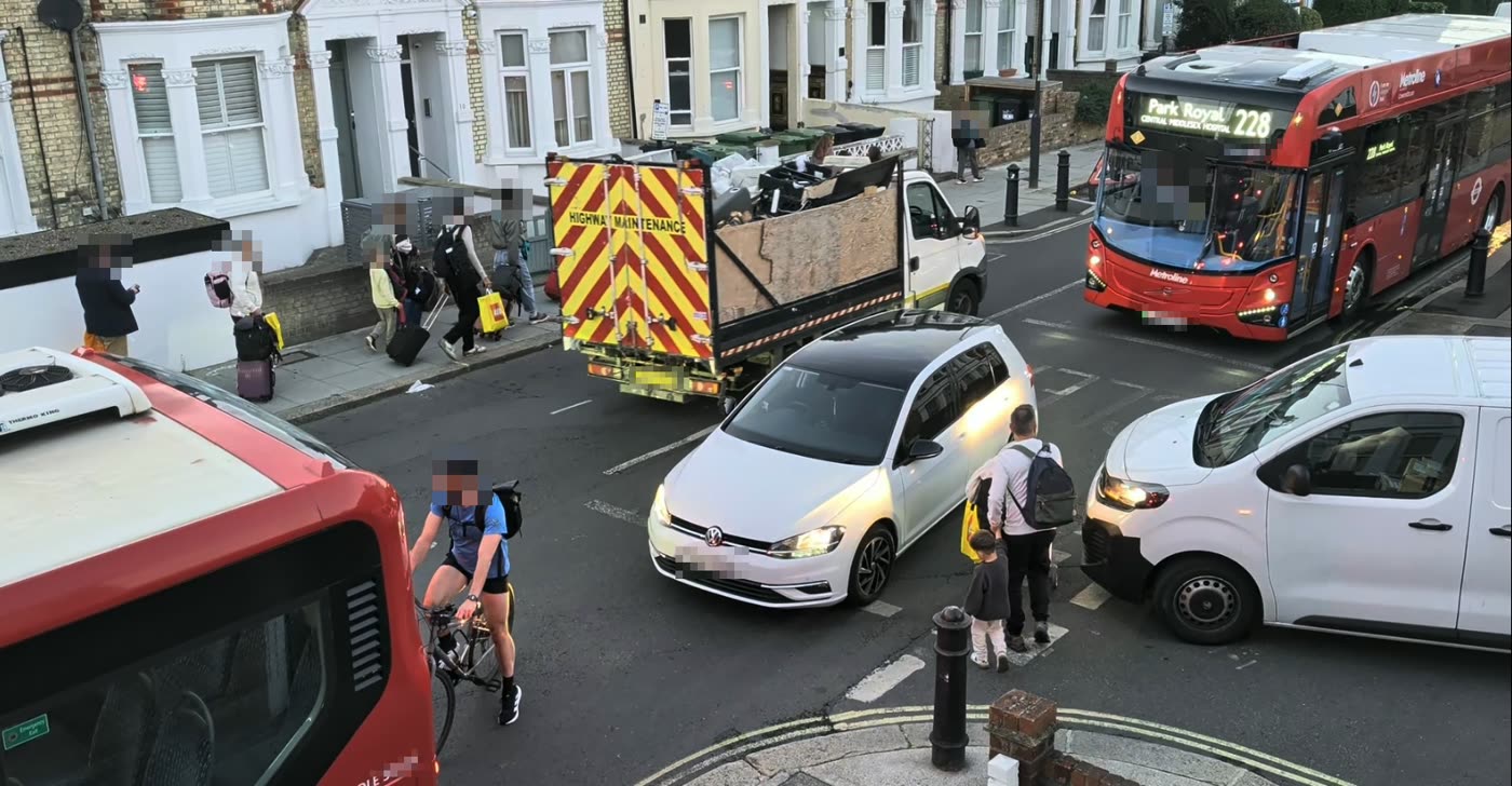 A blocked junction with two buses, a lorry, a car and a van. A cyclist, a parent and a young child pick their way between the vehicles.