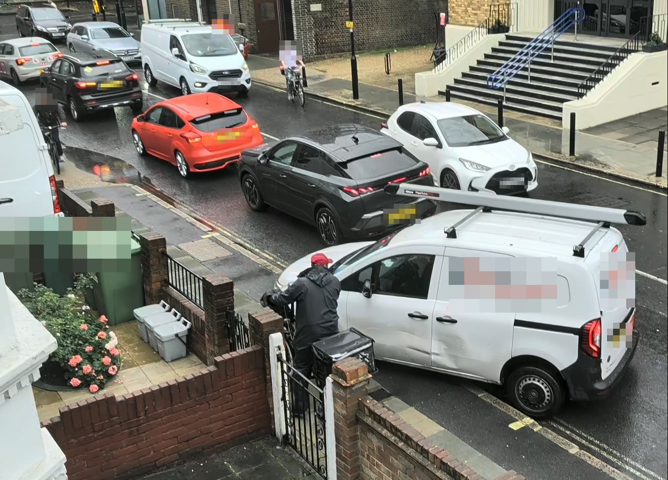 A white van with two wheels on the pavement, squeezing past a queue of cars on a wet day.